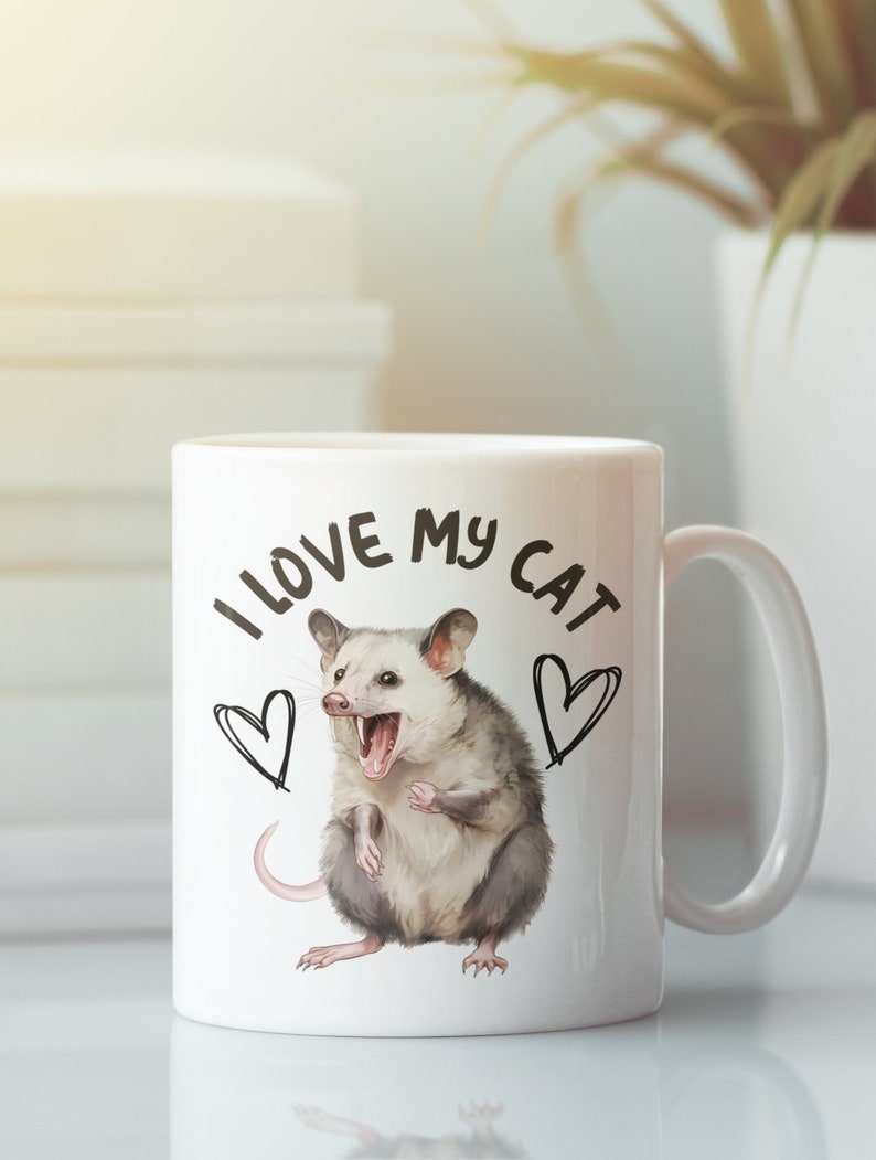 Puede incluir: Taza de cer&aacute;mica blanca con una ilustraci&oacute;n en blanco y negro de una zarig&uuml;eya con el texto "Me encanta mi gato" y dos corazones.