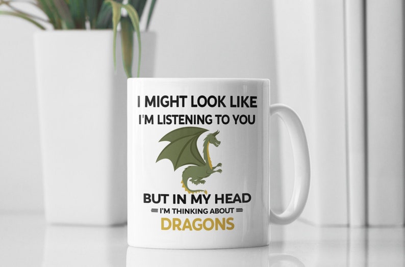 Puede incluir: Taza de cer&aacute;mica blanca con una ilustraci&oacute;n de drag&oacute;n verde y el texto "I might look like I'm listening to you but in my head I'm thinking about DRAGONS".