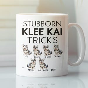 May include: White ceramic coffee mug with a black graphic of a Klee Kai dog breed. The text "Stubborn Klee Kai Tricks" is printed on the mug. The mug has a black dot and the words "Sit, Down, Shake, Come, Fetch, Roll Over, Stay" are printed below the dog graphic.