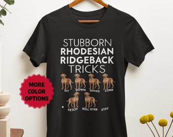 Puede incluir: Camiseta negra con el texto "Stubborn Rhodesian Ridgeback Tricks" e ilustraciones de un perro Rhodesian Ridgeback realizando varios trucos, incluyendo sentarse, tumbarse, dar la pata, venir, traer, rodar y quedarse.