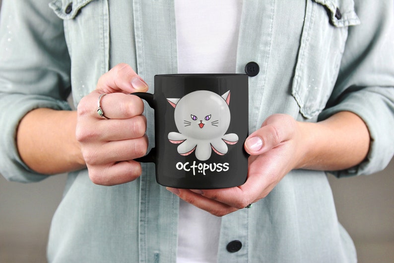 May include: Black ceramic coffee mug with a white octopus with pink accents and cat ears. The octopus has purple eyes and a pink tongue sticking out. The text "OCTOPUSS" is printed in white below the octopus.