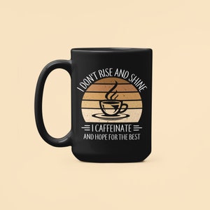 Op de afbeelding: Zwarte koffiemok met een witte afbeelding waarop staat "Ik sta niet op en schijn, ik cafe&iuml;neer en hoop op het beste". De afbeelding toont een retro zonsondergang met een stomende kop koffie.