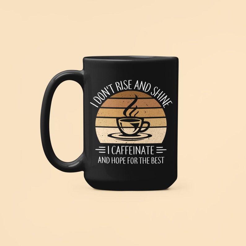 Op de afbeelding: Zwarte koffiemok met een witte afbeelding waarop staat "Ik sta niet op en schijn, ik cafe&iuml;neer en hoop op het beste". De afbeelding toont een retro zonsondergang met een stomende kop koffie.