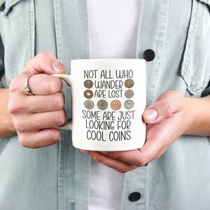 May include: White ceramic mug with a black inscription that reads "Not all who wander are lost, some are just looking for cool coins". The inscription is surrounded by a circle of various coins.