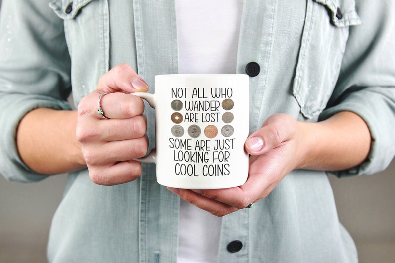 May include: White ceramic mug with a black inscription that reads "Not all who wander are lost, some are just looking for cool coins". The inscription is surrounded by a circle of various coins.
