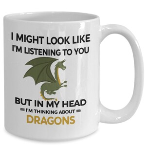 Puede incluir: Taza de cer&aacute;mica blanca con una ilustraci&oacute;n de drag&oacute;n verde y el texto "I might look like I'm listening to you but in my head = I'm thinking about = DRAGONS"