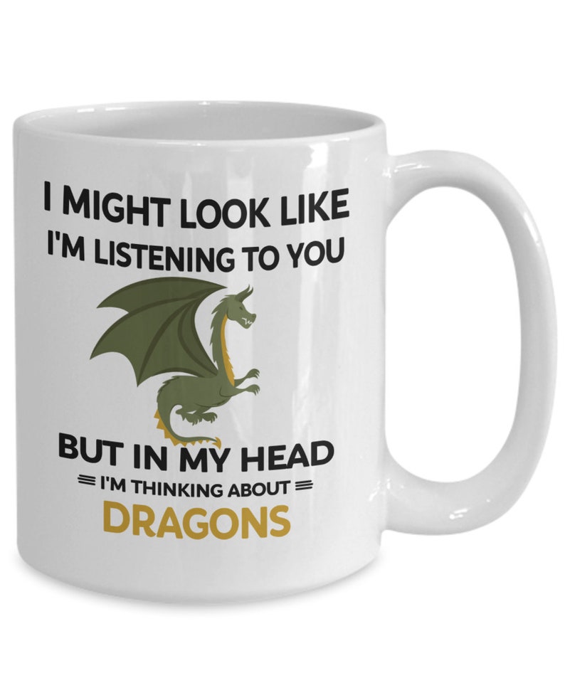 Puede incluir: Taza de cer&aacute;mica blanca con una ilustraci&oacute;n de drag&oacute;n verde y el texto "I might look like I'm listening to you but in my head = I'm thinking about = DRAGONS"