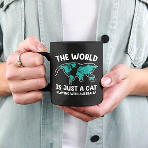 May include: Black ceramic mug with the text "THE WORLD IS JUST A CAT PLAYING WITH AUSTRALIA" and a cat-shaped world map in teal. The mug has a black handle and is held by a person.