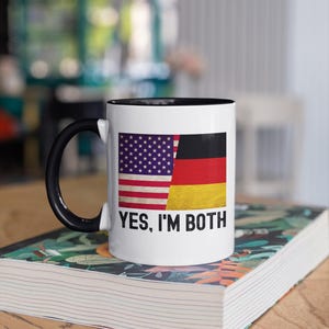 May include: White ceramic mug with a black rim and handle. The mug features an image of the American and German flags side-by-side, with the text "YES, I'M BOTH" printed below the flags.