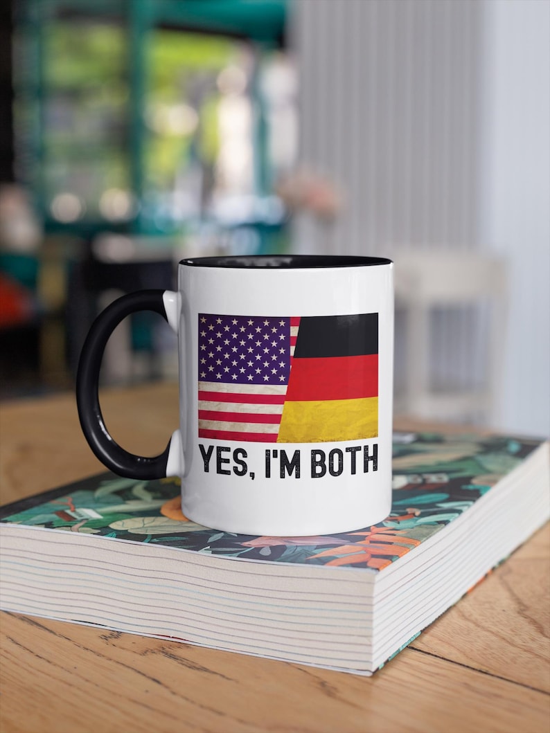 May include: White ceramic mug with a black rim and handle. The mug features an image of the American and German flags side-by-side, with the text "YES, I'M BOTH" printed below the flags.