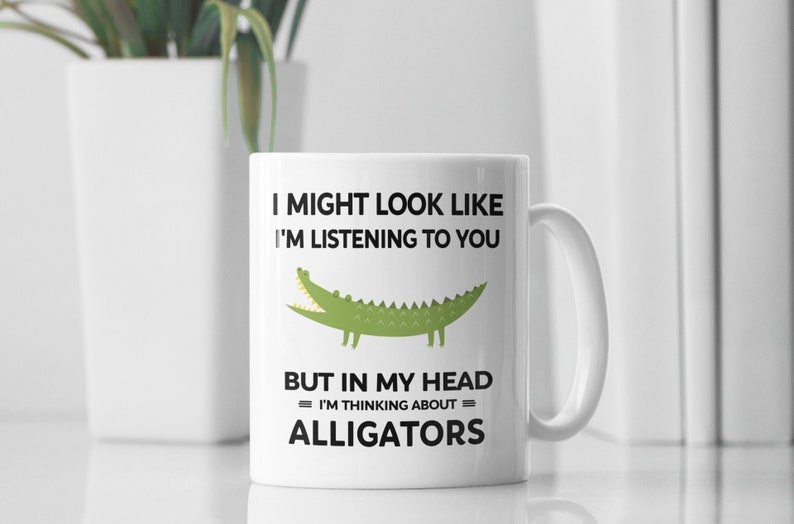 K&ouml;nnte beinhalten: Eine wei&szlig;e Keramiktasse mit einer schwarzen und gr&uuml;nen Alligatorgrafik und dem Text "I might look like I'm listening to you but in my head I'm thinking about = ALLIGATORS =."