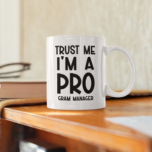 May include: White ceramic mug with black text that reads "Trust me I'm a pro Gram Manager".