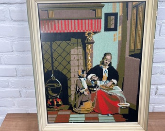 May include: A framed needlepoint artwork depicting a woman in a red dress sitting by a fireplace with a child. The artwork is in a light wood frame with a white mat.