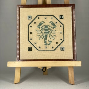 以下が含まれることがあります： 幾何学模様の中に緑色のサソリが描かれたクロスステッチ刺繍の額縁。フレームは茶色の木で、クリーム色のマットが入っています。