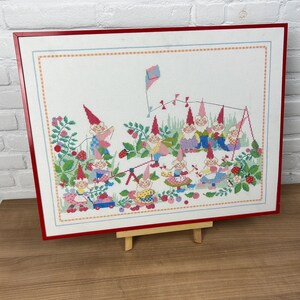 以下が含まれることがあります： 赤いフレームと木製イーゼルが付いた、庭で遊ぶノームのグループを描いたクロスステッチ刺繍の額縁です。