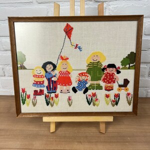 以下が含まれることがあります： 凧揚げをしている子供たちのグループを描いた、額縁に入ったクロスステッチ刺繍。子供たちはカラフルな服を着ていて、笑顔をしています。背景は赤いチューリップのある緑の草原です。