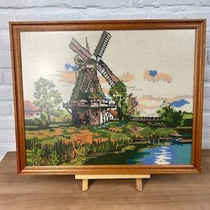 以下が含まれることがあります： オランダの風車を描いた額入りのクロスステッチアート。緑と茶色のディテールが施された風車、木々、水面、カラフルな風景が特徴です。フレームは木製です。