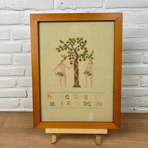 以下が含まれることがあります： エデンの園を描いた額縁付きのクロスステッチ刺繍。シーンには赤いリンゴがなった木、ヘビ、木の両側に立っている2人の人物が描かれている。アルファベットはシーンの下に刺繍されている。