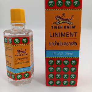 May include: A clear glass bottle of Tiger Balm Liniment with a yellow cap and its matching rectangular orange and red box. Both items feature the Tiger Balm logo, the text 1 FL. OZ. 28ml, and green circular icons with Chinese characters.