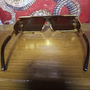 May include: Gold-framed sunglasses with brown lenses. The sunglasses have a geometric design with a rectangular shape. The temples are black and curve around the ears. The background features a red and white snake design.
