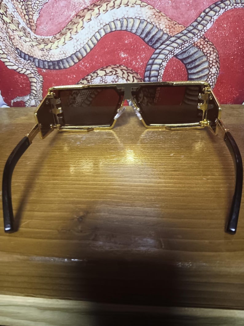 May include: Gold-framed sunglasses with brown lenses. The sunglasses have a geometric design with a rectangular shape. The temples are black and curve around the ears. The background features a red and white snake design.