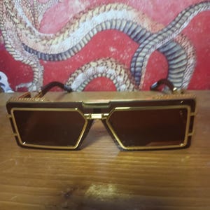 May include: Sunglasses with a gold frame and dark brown lenses. The sunglasses have a rectangular shape with a geometric design. The background features a red wall with a dragon illustration.