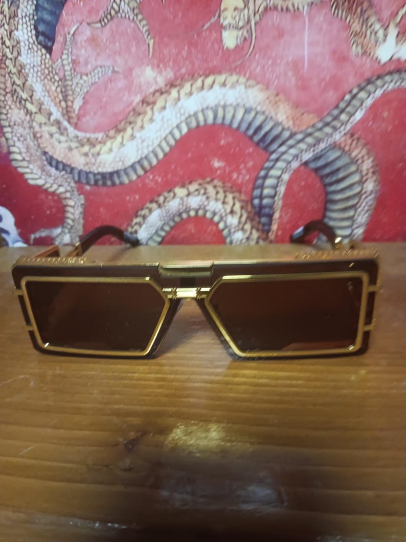 May include: Sunglasses with a gold frame and dark brown lenses. The sunglasses have a rectangular shape with a geometric design. The background features a red wall with a dragon illustration.