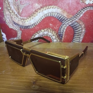 May include: Rectangular sunglasses with brown lenses and gold-coloured frames. The sunglasses have a geometric design. The temples are dark brown. The background features a red wall with a dragon illustration.
