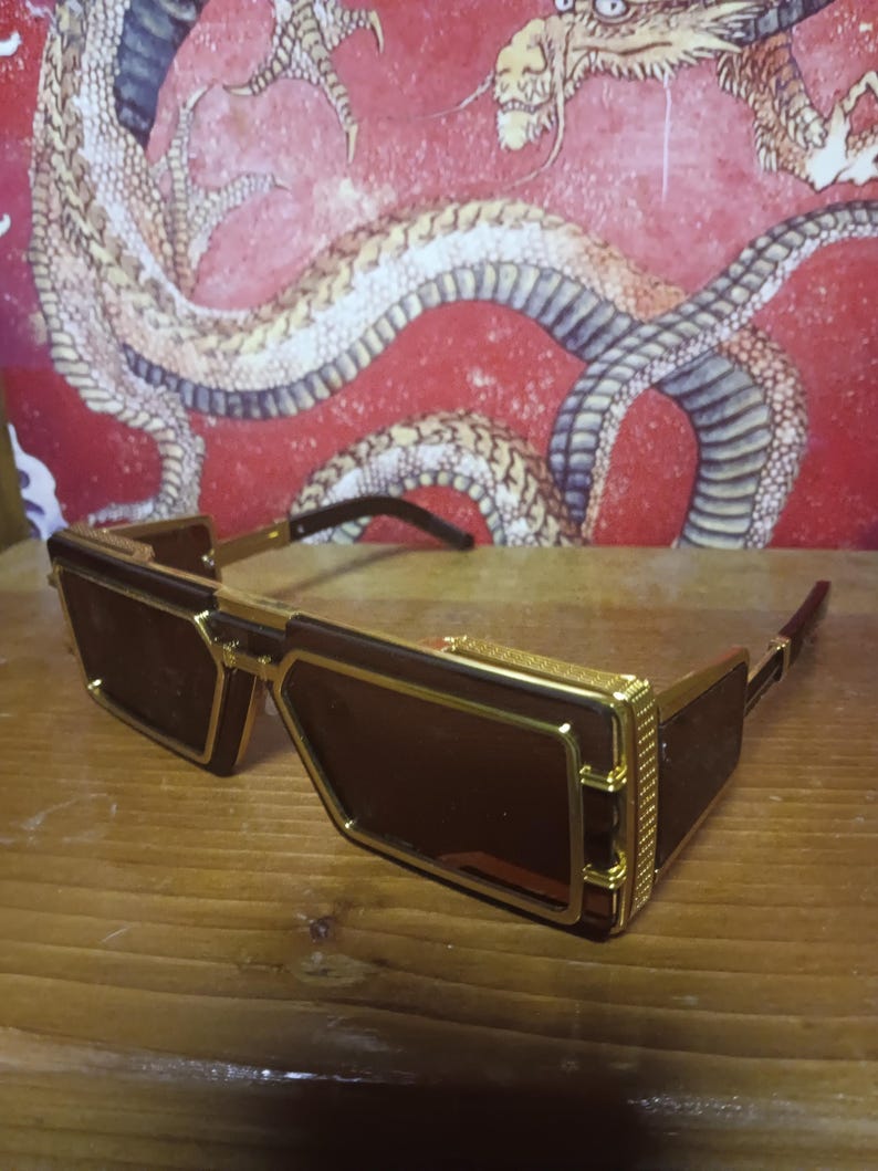 May include: Rectangular sunglasses with brown lenses and gold-coloured frames. The sunglasses have a geometric design. The temples are dark brown. The background features a red wall with a dragon illustration.