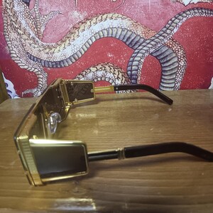 May include: Gold-framed sunglasses with dark lenses and black arms. The sunglasses have a rectangular shape and a decorative pattern on the frame. A red wall with a dragon illustration forms the background.