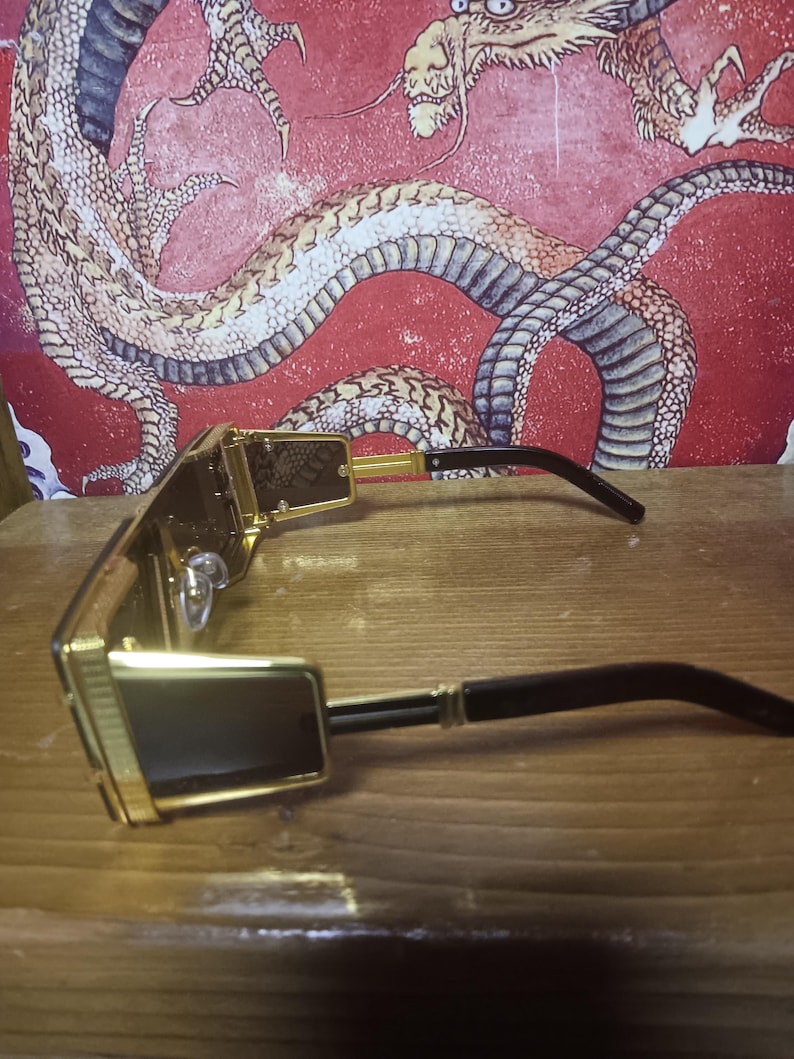 May include: Gold-framed sunglasses with dark lenses and black arms. The sunglasses have a rectangular shape and a decorative pattern on the frame. A red wall with a dragon illustration forms the background.