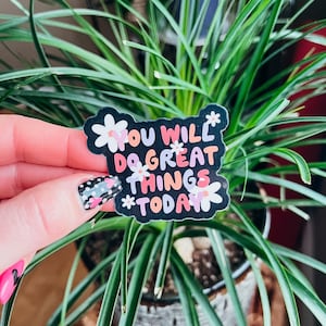 Puede incluir: Una pegatina negra con el texto "YOU WILL DO GREAT THINGS TODAY" en blanco, naranja y rosa, con detalles de flores blancas. La pegatina se sostiene frente a una planta verde.