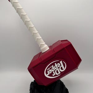 May include: A novelty hammer in the shape of a Dr. Pepper can. The hammer is primarily burgundy with a white handle and the Dr. Pepper logo. It rests on a black, textured base.