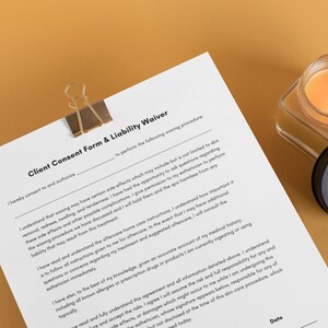 May include: A white client consent form and liability waiver with the text "Client Consent Form & Liability Waiver" at the top. The form is on a light brown surface with a lit candle and a black lid with a gold design in the background.