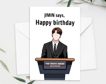 May include: A white greeting card with the text "JIMIN says, Happy birthday" in black. The card features an illustration of a person in a black suit standing behind a podium with "THE WHITE HOUSE WASHINGTON" printed on it. An envelope is on the left.