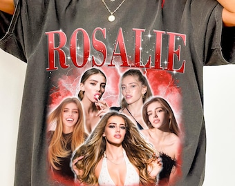 以下が含まれることがあります： 上部に大きな赤い文字でROSALIEと書かれたチャコールグレーのTシャツ。テキストの下には、長い茶色の髪をした同じ人物の5つのポートレートのグラフィックコラージュがあり、稲妻の効果が施された赤い背景に配置されています。