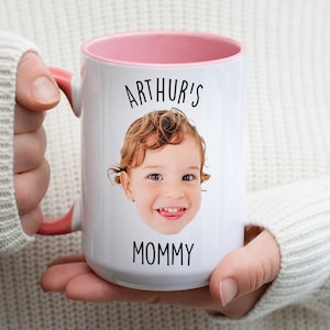 May include: White ceramic mug with a pink interior and handle. The mug features a smiling child's face and the text "ARTHUR'S MOMMY" in black. The mug is held by a person wearing a white sweater.