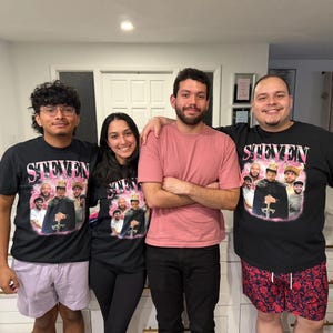 May include: Two black t-shirts featuring a pink graphic print with the name STEVEN in bold letters above a collage of photos of a person wearing a crown and holding a sword, surrounded by smaller portraits of various individuals in a group setting.
