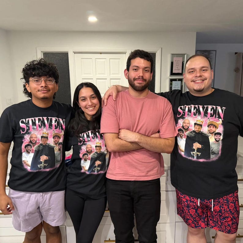 May include: Two black t-shirts featuring a pink graphic print with the name STEVEN in bold letters above a collage of photos of a person wearing a crown and holding a sword, surrounded by smaller portraits of various individuals in a group setting.