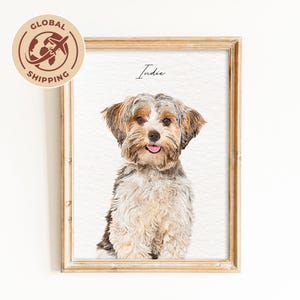 May include: A framed portrait of a small dog with brown, black, and white fur, featuring the name Indie written in black script above the dog's head. The artwork is set within a light-toned wooden frame against a plain white background.