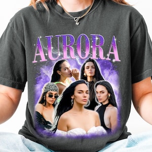 Peut inclure: Un t-shirt gris anthracite à manches courtes avec une impression graphique comportant le mot AURORA en grandes lettres violettes. Le motif inclut quatre portraits photographiques de la même personne sur un fond d'éclairs et de nuages violets.