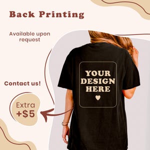 May include: A black t-shirt featuring a rectangular graphic on the back with the text "YOUR DESIGN HERE" in a beige, wavy font, accompanied by a small heart icon. The design is centered on the back of the shirt, which is shown from behind.