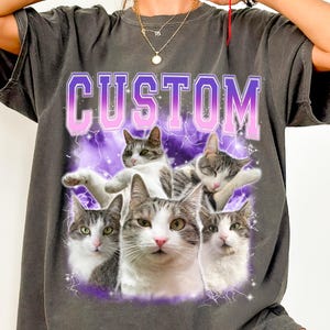May include: A charcoal gray t-shirt featuring the word CUSTOM in large purple and white block letters at the top. Below the text, a graphic design displays five different poses of a gray and white tabby cat against a purple lightning background.