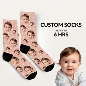 May include: Two custom socks with a repeating pattern of a baby's face and small white hearts on a light pink background. The socks have black heels and toes. Text on the right says "CUSTOM SOCKS READY IN 6 HRS".