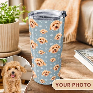 May include: A cylindrical stainless steel tumbler with a light blue background featuring a repeating pattern of small white dog paw prints, bones, and the face of a light brown fluffy dog with a pink tongue visible in its open mouth.