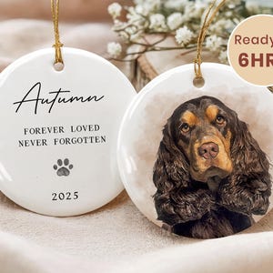 Puede incluir: Dos adornos navideños de cerámica blanca. Uno presenta un retrato de perro con pelaje marrón y canela. El otro adorno tiene el nombre "Autumn", el texto "Forever Loved, Never Forgotten", una huella de pata y el año 2025. Ambos tienen cuerda dorada.