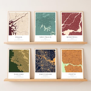 Może przedstawiać: Sześć oprawionych wydruków map miast, eksponowanych na drewnianych półkach. Mapy przedstawiają miasta takie jak Osaka, São Paulo, Montreal, Oakland, Amsterdam i Porto. Każda mapa ma unikalną kolorystykę, z nazwami miast i współrzędnymi na dole.