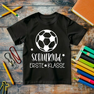 May include: Black t-shirt with a white soccer ball graphic and the text "SCHULKIND ERSTE KLASSE" in white.