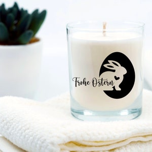 May include: A white candle in a clear glass jar with a black and white sticker featuring a bunny silhouette and the text "Frohe Ostern" (Happy Easter).