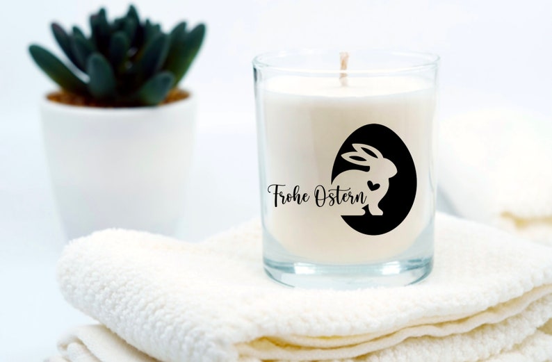 May include: A white candle in a clear glass jar with a black and white sticker featuring a bunny silhouette and the text "Frohe Ostern" (Happy Easter).